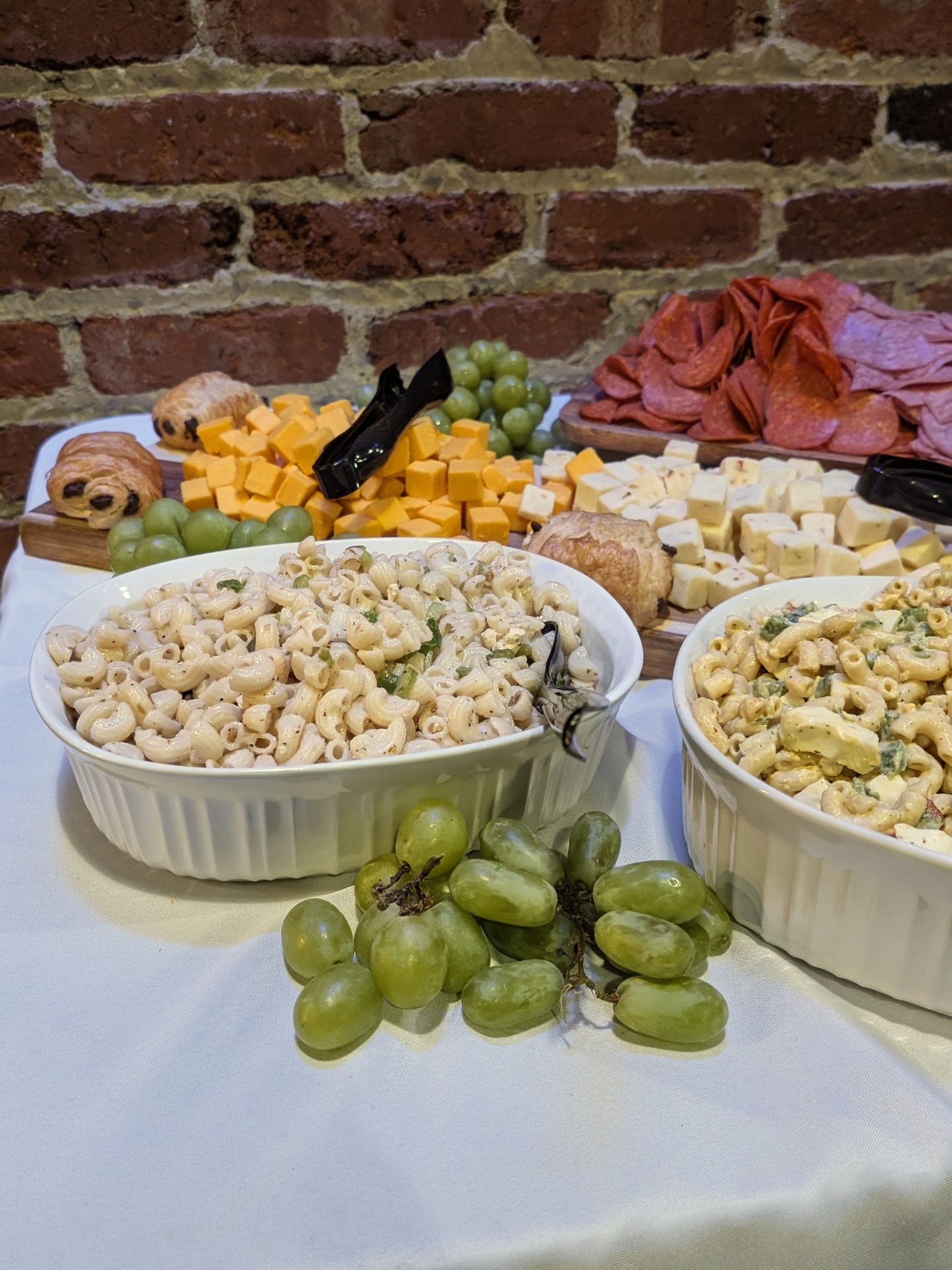Two pasta salad bowls — classic mayo macaroni and an Italian-style with peppers — beside the cheese board and grapes